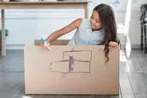 Free photo smiling girl sitting inside the cardboard box drawing with marker