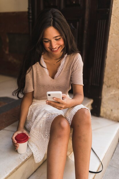 Smiling girl sitting on steps holding disposable coffee cup using cell phone