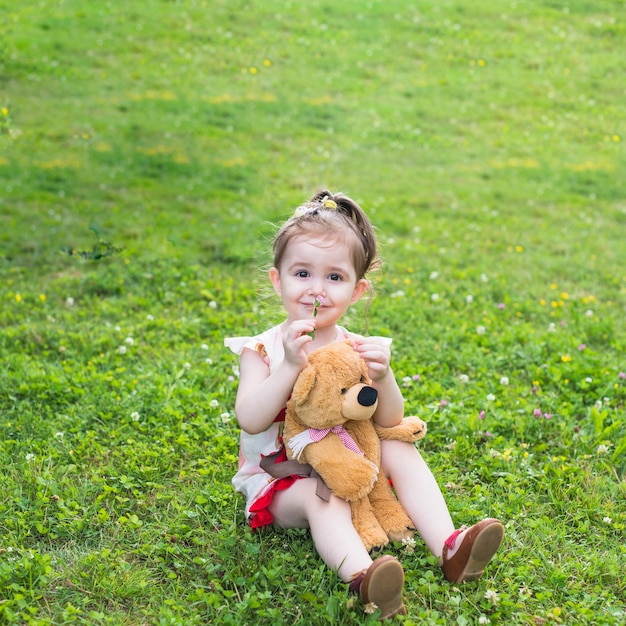 Free Photo smiling girl sitting with teddy bear smelling flower in the park