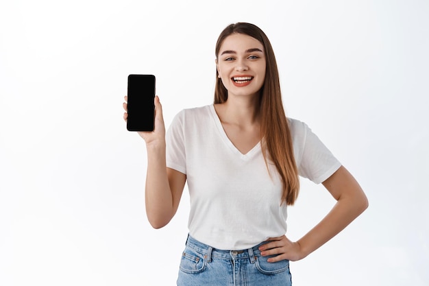 Smiling girl student shows shopping app, empty smartphone screen and looks pleased, recommends online shop or application, stands against white background