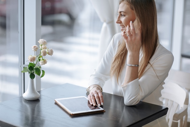 Free photo smiling girl talking on the phone with a tablet on the table