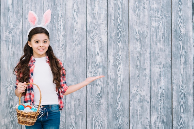 Free photo smiling girl with bunny ears holding basket of easter eggs presenting against gray wooden desk