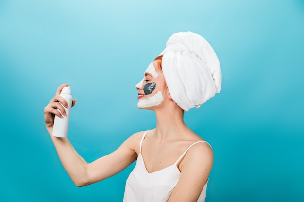 Smiling girl with towel on head holding cosmetics bottle. Studio shot of laughing woman with face mask standing on blue background.