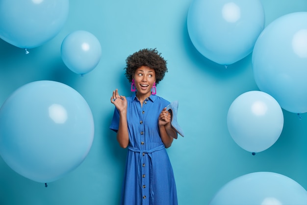 Free photo smiling glad afro american woman chooses outfit for birthday party, holds blue shoes on high heels to fit dress, looks aside happily, poses near inflated balloons flying around. women, clothes