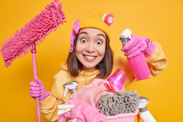 Free Photo smiling glad young asian woman holds spray bottle and mop stands near laundry basket with chemical bottles wears rubber gloves uses house