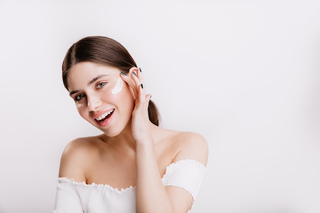 Smiling green-eyed girl puts cream on clean face. Brunette in white top posing on isolated wall.