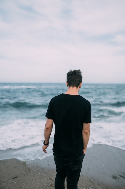 Smiling guy in a black T-shirt stands on the sandy seashore.