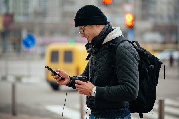 Smiling guy in eyeglasses with headset, using his smartphone, holding a cup of hot coffee