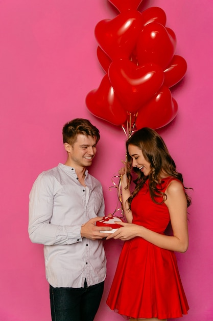 Smiling guy gives box with present, red balloons in form of heart for his beautiful girlfriend