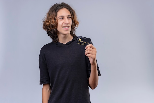 Free Photo smiling guy with long hair in black t-shirt holding a bank card on white wall