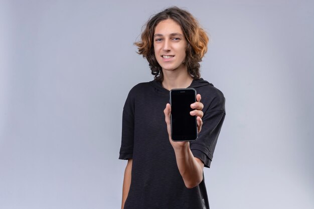 Smiling guy with long hair in black t-shirt holding a phone on white wall