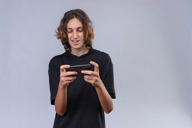 Smiling guy with long hair in black t-shirt playing game from a phone on white wall