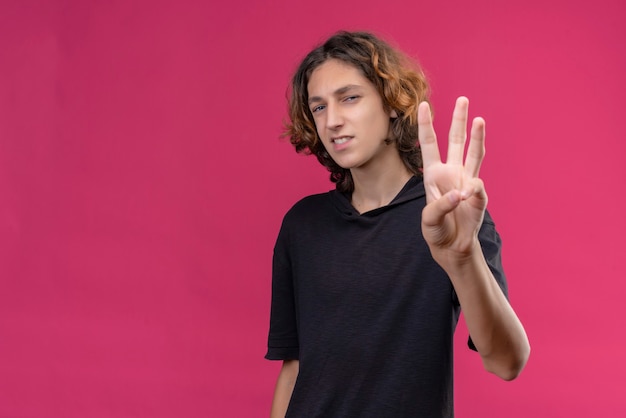 Free photo smiling guy with long hair in black t-shirt showimg three with his fingers on pink wall