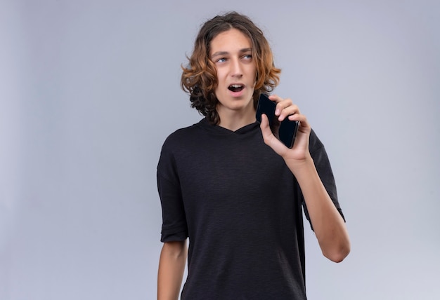 Smiling guy with long hair in black t-shirt speak on phone through speaker on white wall