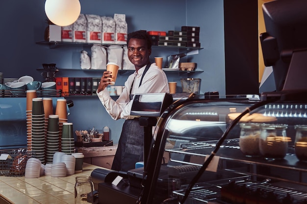 Free Photo smiling handsome african american barista in uniform holds cup of coffee while standing in his coffee shop.