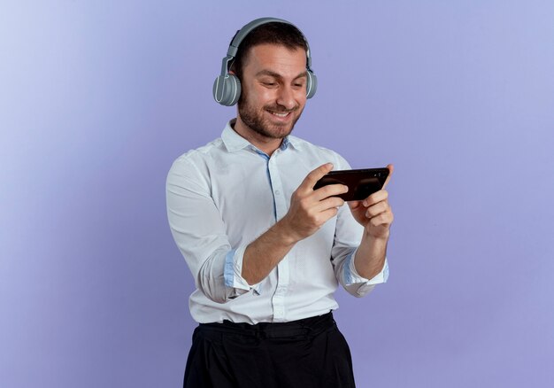 Smiling handsome man on headphones holds and looks at phone isolated on purple wall