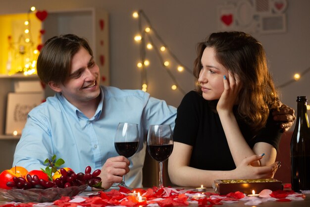 Smiling handsome man holding glass of wine and looking at pleased pretty woman sitting at table in living room on valentine's day