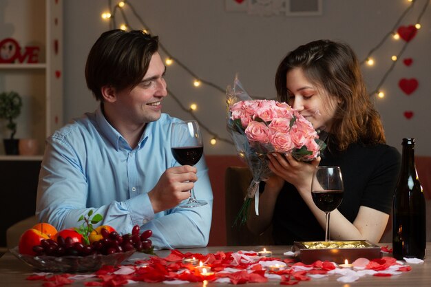 Smiling handsome man holding glass of wine and looking at pretty woman sniffing bouquet of flowers sitting at table in living room on valentine's day