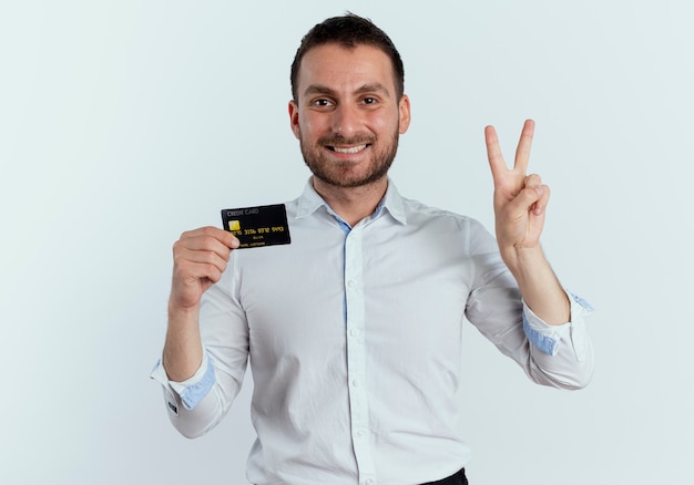 Smiling handsome man holds credit card and gestures two with hand isolated on white wall