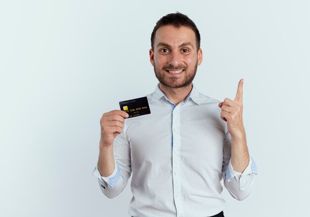 Smiling handsome man holds credit card and points up isolated on white wall