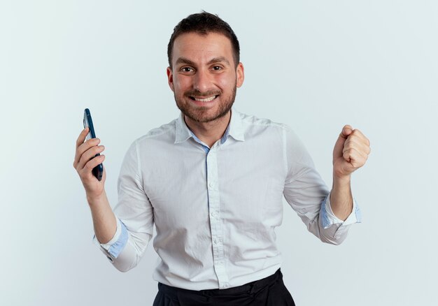 Smiling handsome man holds phone and keeps fist isolated on white wall