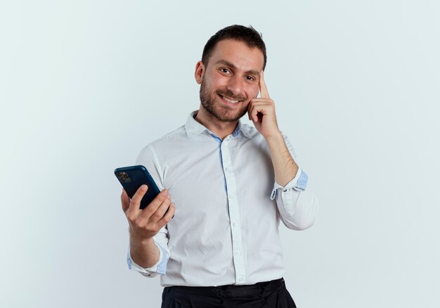 Smiling handsome man puts finger on temple holding phone isolated on white wall