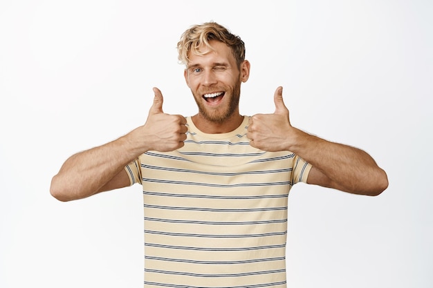 Free Photo smiling handsome man showing thumbs up and winking say yes praise and compliment standing in tshirt over white background