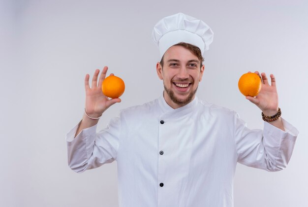 A smiling handsome young bearded chef man wearing white cooker uniform and hat holding oranges while looking on a white wall