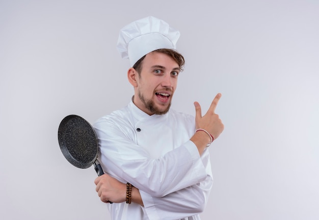 A smiling handsome young bearded chef man wearing white cooker uniform and hat pointing up and holding frying pan while looking on a white wall