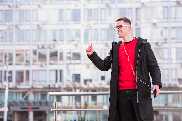 Smiling handsome young man listening music on earphone showing thumb up sign at outdoors