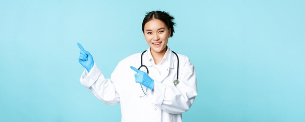 Smiling happy female doctor or nurse pointing fingers left wearing medical uniform and gloves showin