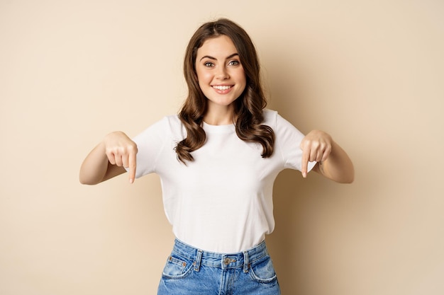 Smiling happy girl pointing fingers down and showing banner, company logo below, click link, standing over beige background