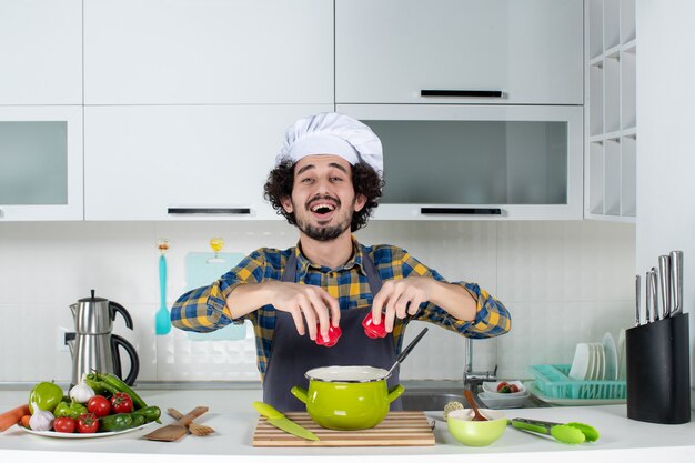 Smiling and happy male chef with fresh vegetables adding red peppers into meal in the white kitchen