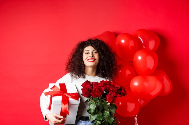 Smiling happy woman holding box with gift and red roses from boyfriend celebrating valentines day st...