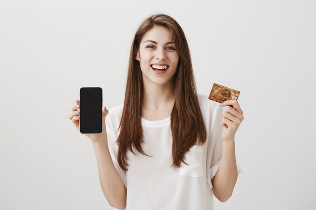 Smiling happy woman showing mobile phone display and credit card. Promo of shopping application