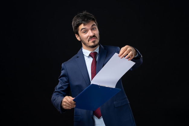 Free photo smiling and happy young adult in suit holding documents and checking statistics in it on isolated dark wall