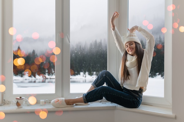 Free photo smiling happy young attractive woman in stylish white knitted sweater, scarf and hat sitting at home on windowsill at christmas having fun holding hands up, winter forest background view, lights bokeh