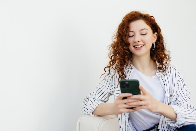 Free Photo smiling happy young woman with red hair texting on phone lying on the couch at home