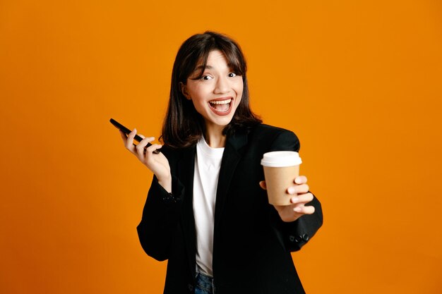 Smiling holding coffee cup with phone young beautiful female wearing black jacket isolated on orange background