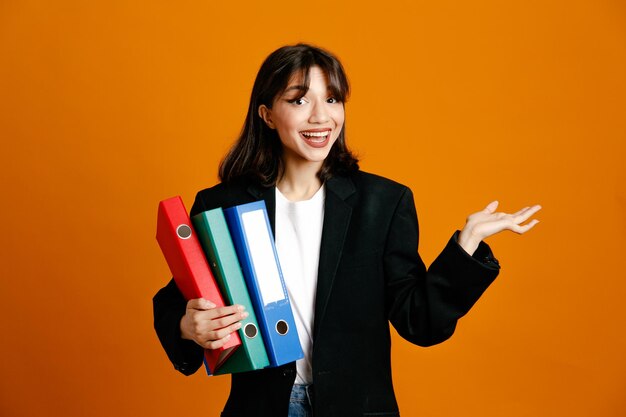 Smiling holding folders young beautiful female wearing black jacket isolated on orange background