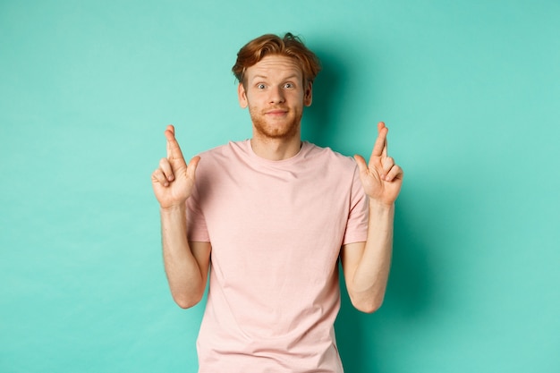 Free photo smiling hopeful man with red hair making a wish, cross fingers for good luck and expecting something good, standing over turquoise background