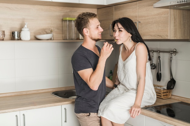 Free Photo smiling husband feeding food to his wife sitting on kitchen counter