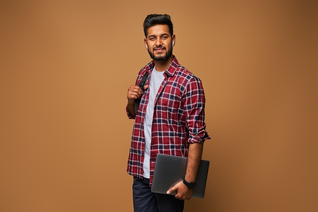 Free photo smiling indian man in casual close with laptop and backpack on pastel wall