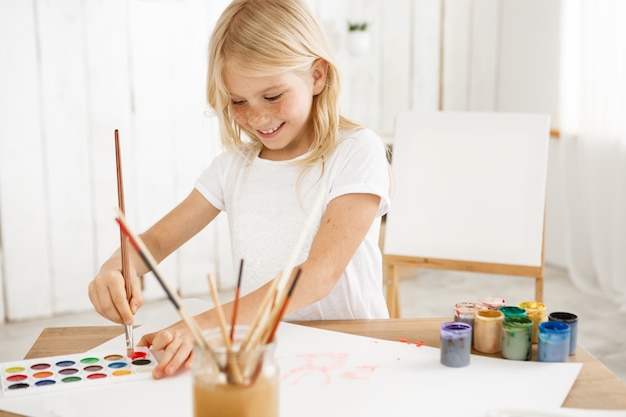 Smiling, inspired girl with blonde hair and freckles joyfully deeping brush into red paint, having new idea for a picture.