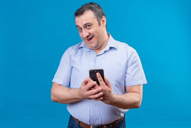 Smiling and joyful man in blue striped shirt holding his mobile phone on a blue background