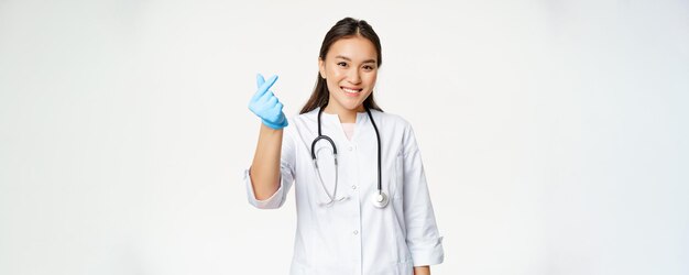 Smiling korean female doctor shows finger heart gesture in rubber gloves wearing medical uniform looking happy at camera white background