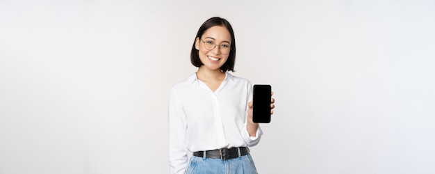 Free photo smiling korean woman showing smartphone screen demonstrating mobile application standing over white