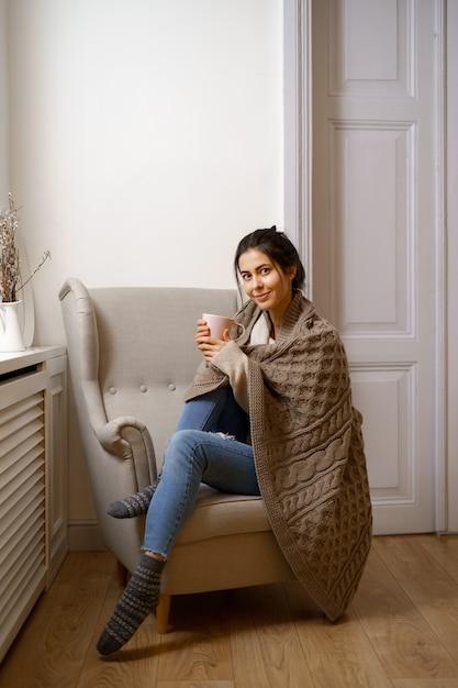 Smiling lady in smart trendy wear is sitting on armchair with a cup of tea.