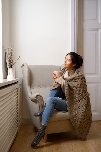 Smiling lady in smart trendy wear is sitting on armchair with a cup of tea.