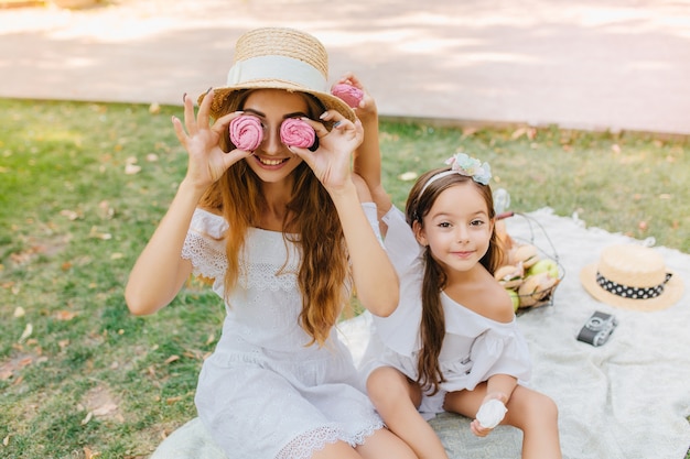 Smiling lady in white dress holding pink gingerbreads like glasses, sitting on blanket with daughter. Pretty little girl with ribbon posing next to joking mother during picnic.